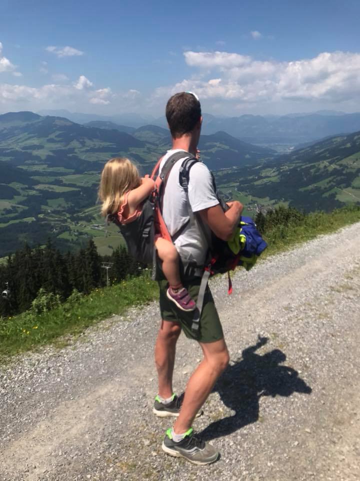 En man går på en grusväg med ett barn i en ryggsäck och njuter av skönheten på sin sommarsemester i Westendorf. De ser ut över ett naturskönt landskap med gröna kullar och avlägsna berg under en blå himmel med spridda moln medan han håller några redskap i handen.