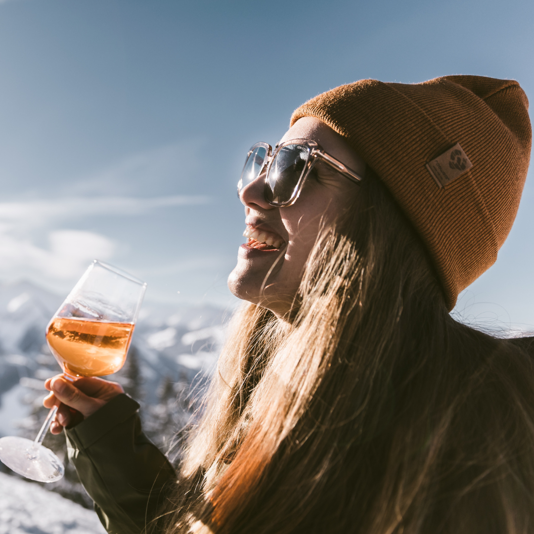 Destinationer för skidsemester i Österrike En person i brun mössa och solglasögon ler medan han håller i ett glas vin utomhus. I bakgrunden syns snötäckta berg och en klarblå himmel som fångar den livliga afterski-atmosfären.