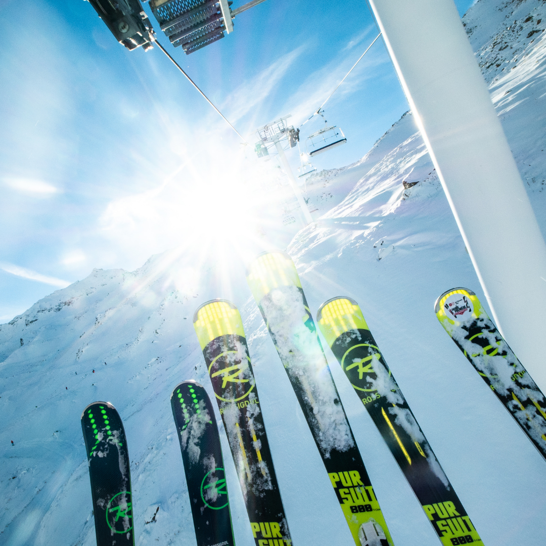 Närbild av flera par skidor som pekar uppåt under en solig, klarblå himmel. En skidlift syns ovanför, med snötäckta berg i bakgrunden. Solljuset skapar ett starkt sken som framhäver det vintriga landskapet - perfekt för att fira nyår på skidor. 