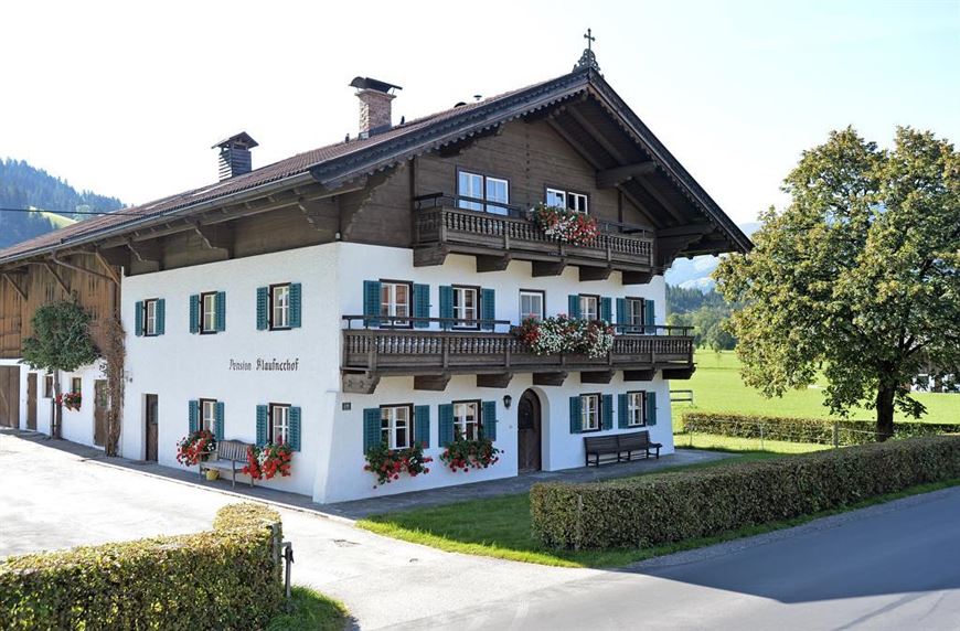 Pension Klausnerhof ligger på en lugn gata och är ett traditionellt alphus med en träbalkong som pryds av röda blommor. Den vita fasaden har gröna fönsterluckor. En frodig grön äng och träd syns under en klarblå himmel. 