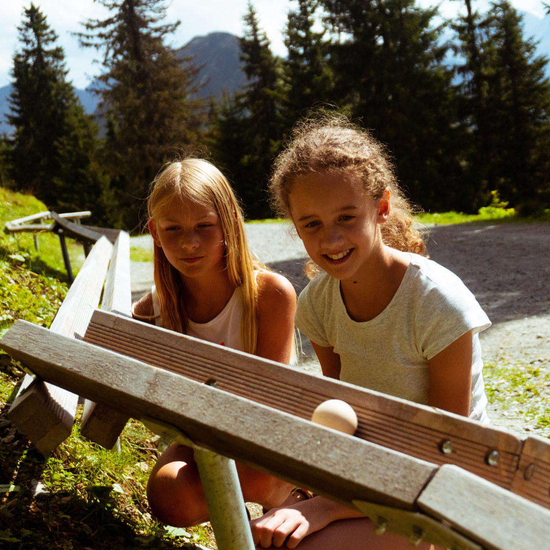 To piger er udendørs under deres sommerferie i Westendorf og leger med en lille trærampe og en hvid bold. De er smilende og fokuserede, omgivet af solbeskinnede træer og de majestætiske bjerge i baggrunden. Scenen er levende og grøn og fanger glæden ved deres ferieeventyr.