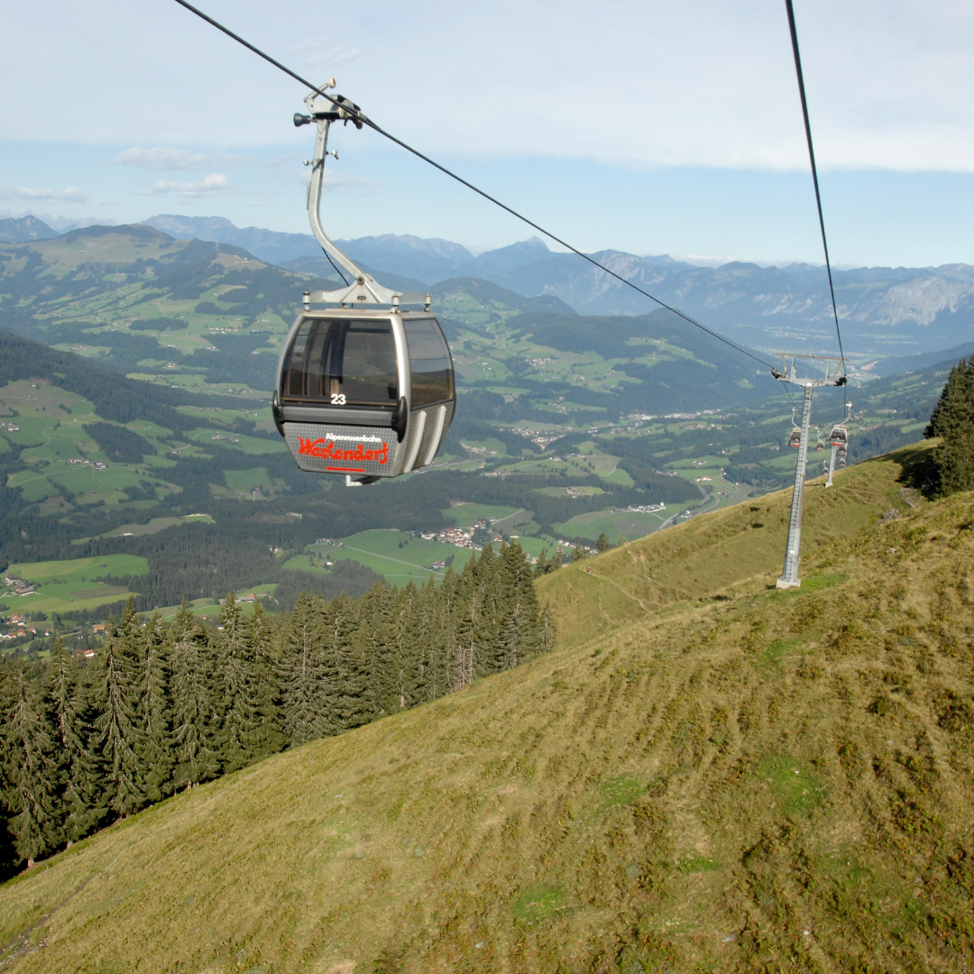 En gondollift stiger op ad en bjergside i Westendorf under sommerferien, med grønne marker og skove, der breder sig nedenfor. Adskillige andre gondoler følger kabellinjen, mens bølgende bakker og fjerne bjerge soler sig under en klar blå himmel.