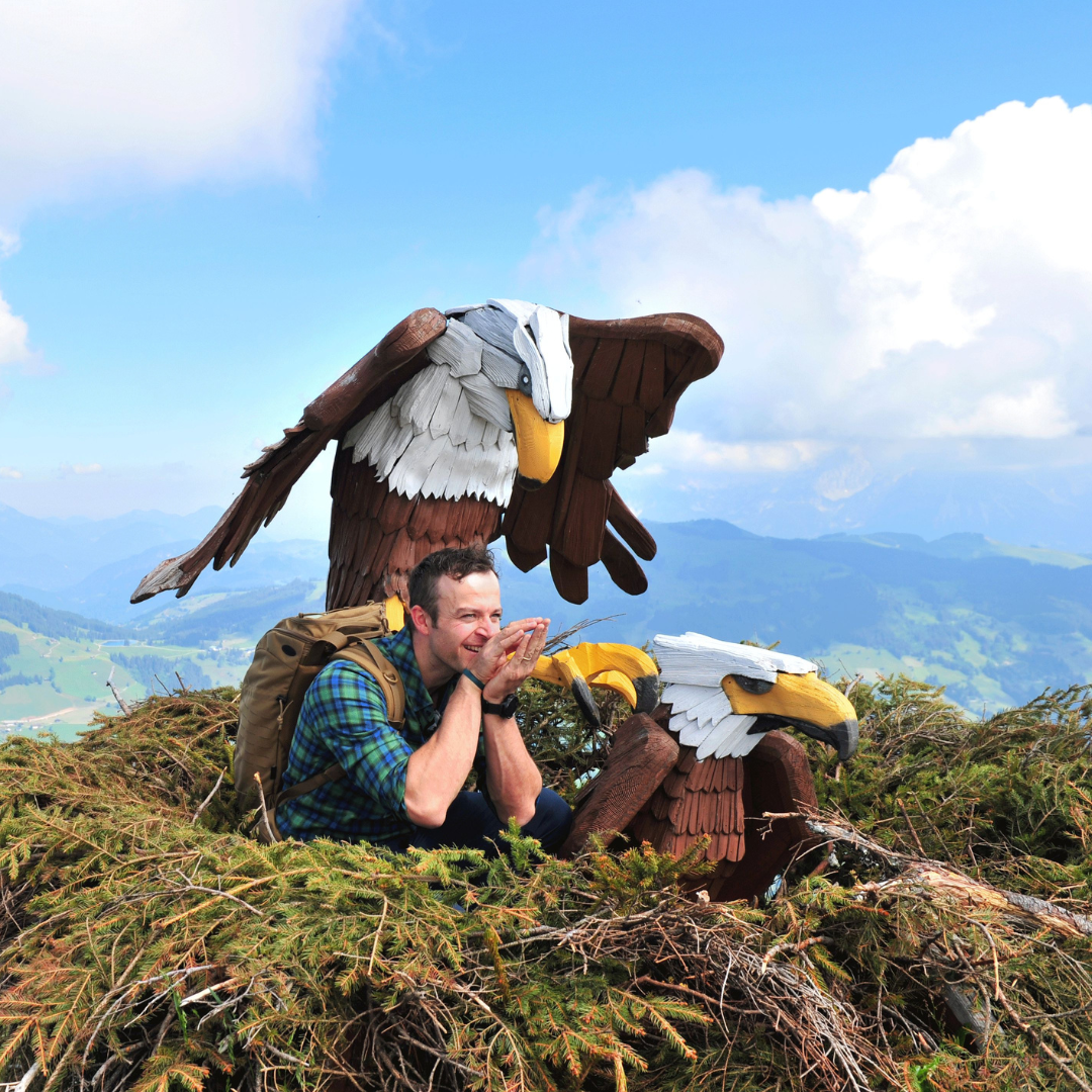 En person i en plaidskjorte sidder i en stor rede på et bjerg, omfavnet af to ørneskulpturer i træ. Det er de perfekte rammer for en "Sommerferie i Westendorf", med klar blå himmel og grønne bakker, der maler en idyllisk kulisse.