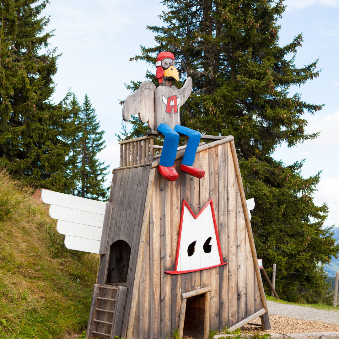En finurlig træstruktur, der ligner en kylling, med en pilotfigur på toppen, bringer et strejf af Sommerferie i Westendorf. Med hvide vinger og et tegneserielignende ansigt nær indgangen står den midt blandt grønne træer og græs, indrammet af en delvist overskyet himmel.
