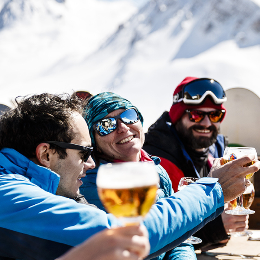 Tre personer i vinterudstyr skåler med glas øl i udendørs sneklædte bjerge og omfavner afterski-stemningen. De smiler og ser ud til at nyde den solrige dag og det smukke landskab med snedækkede tinder synlige i baggrunden.