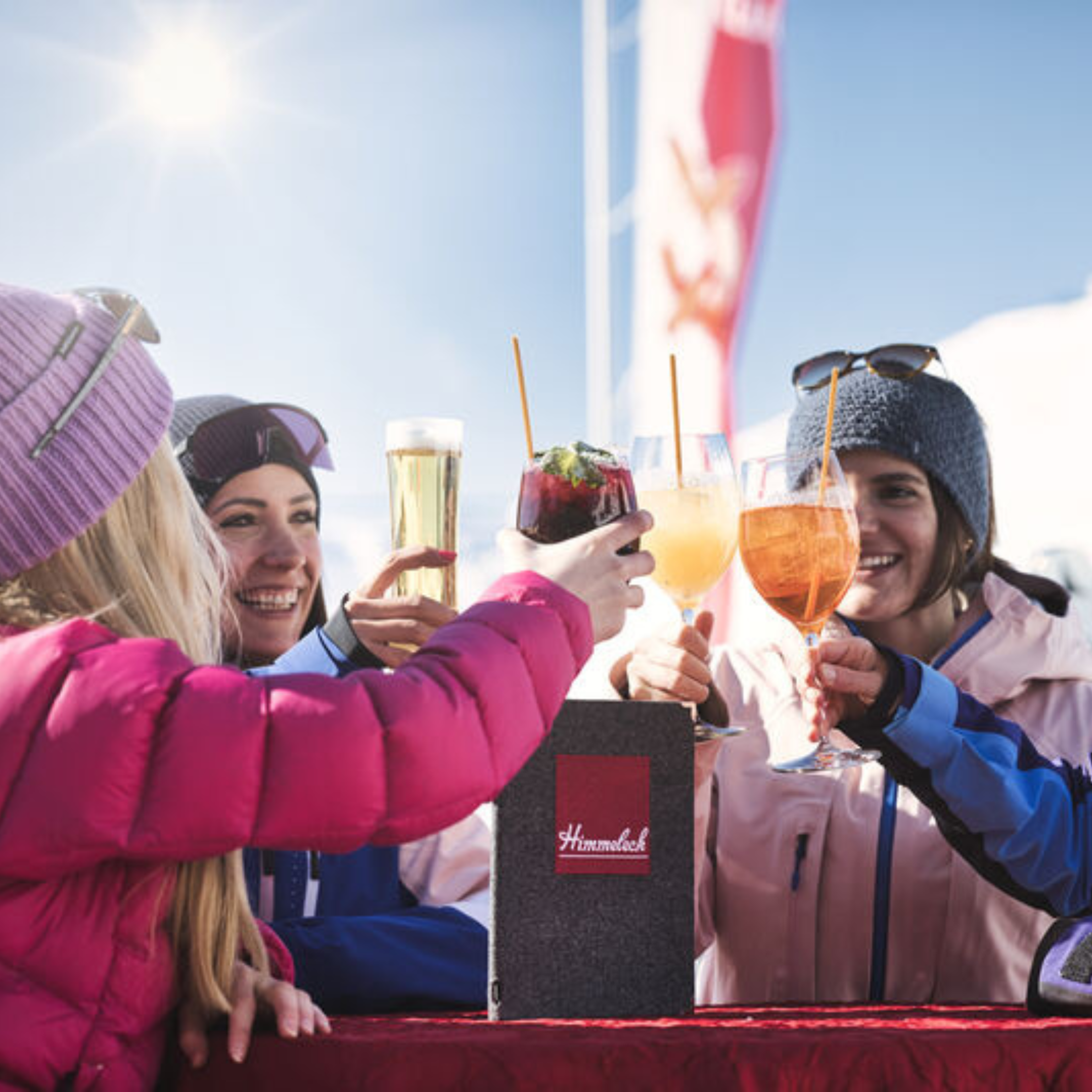 Fire personer i vintertøj skåler med forskellige drinks og fanger den sande essens af afterski udendørs. De smiler, mens de sidder ved et bord med snedækket kulisse, mens sollyset fremhæver deres muntre sammenkomst.