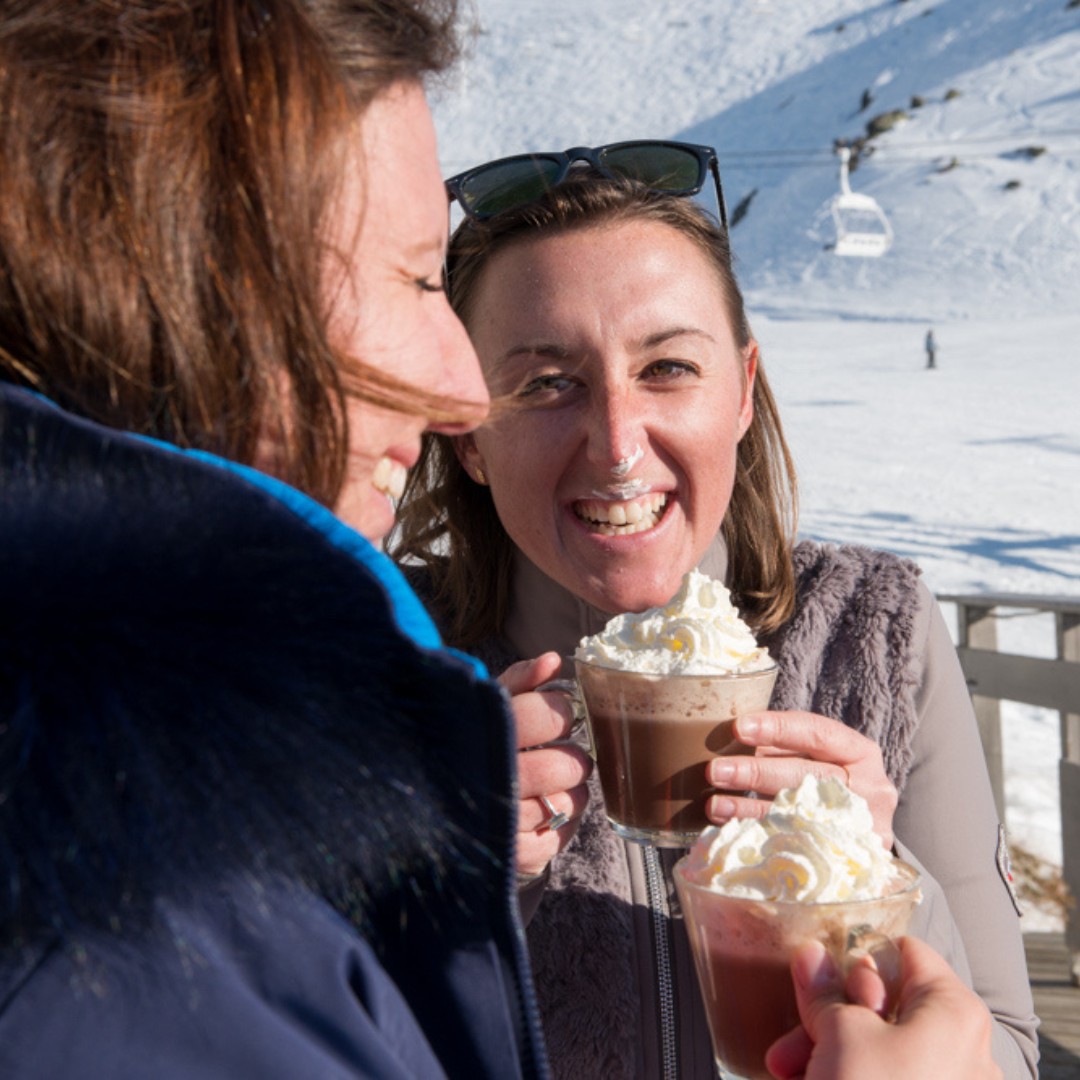 To mennesker, der smiler og holder krus med varm chokolade toppet med flødeskum, nyder en hyggelig afterski-stund. De er klædt varmt på og står udenfor i et snelandskab med en skilift synlig i baggrunden.