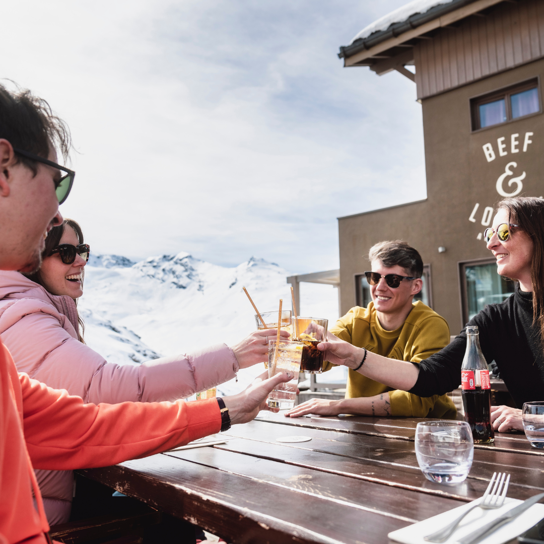 En gruppe på fire personer, pakket ind i varmt tøj, sidder ved et udendørs bord og soler sig i det solrige afterski-skær. De klirrer glade glas, med sneklædte bjerge, der giver en fantastisk baggrund for deres latter og kammeratskab.