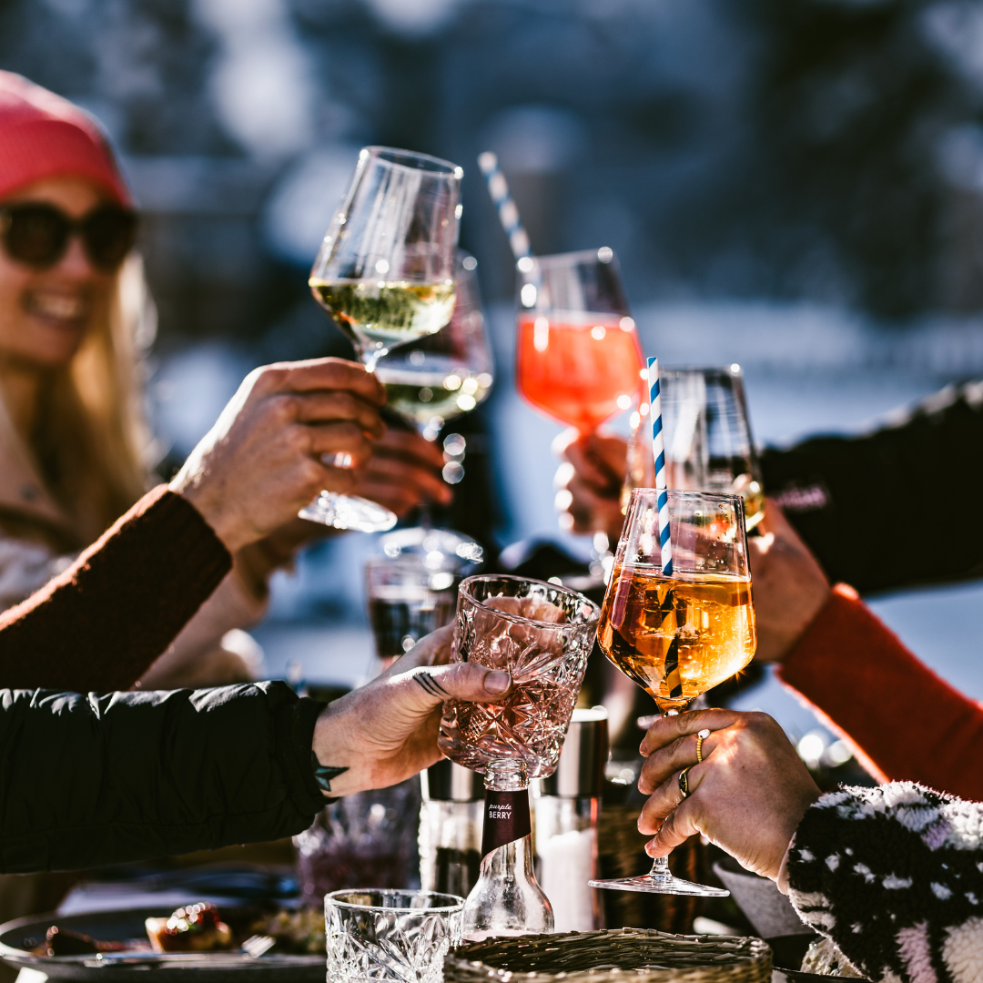 En gruppe mennesker udendørs jubler med forskellige drinks i elegante glas, der fanger essensen af afterski-festligheder. Rammerne fremstår glade og solrige med sne i baggrunden og gruppen bundtet sammen i varmt tøj.