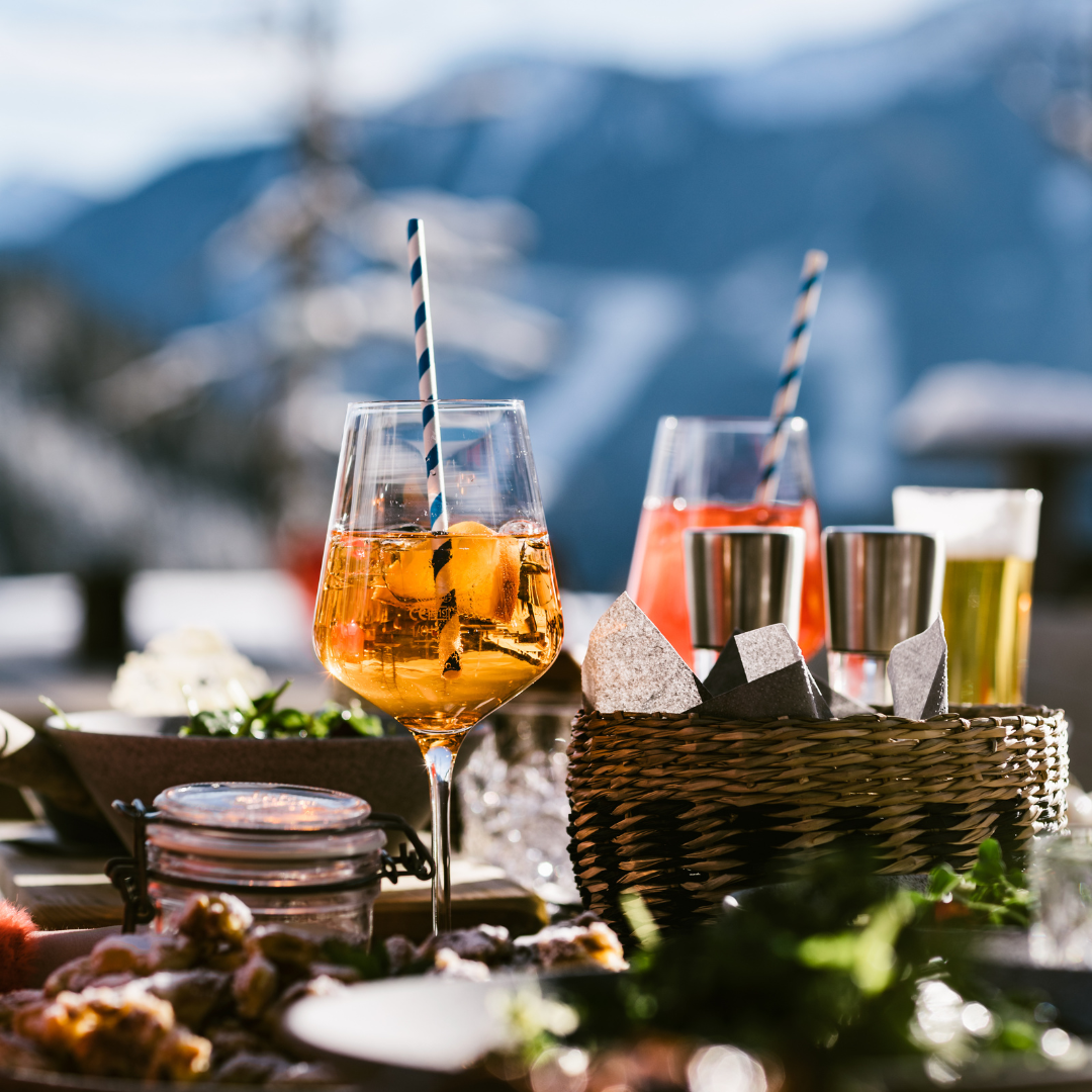 Et bord dækket til udendørs nydelse med to cocktails garneret med stribede sugerør, en kurv med metalkopper og snacks inkarnerer perfekt afterski-stemningen. Sneklædte bjerge og en klar himmel danner en fantastisk kulisse, der skaber en hyggelig, festlig atmosfære.