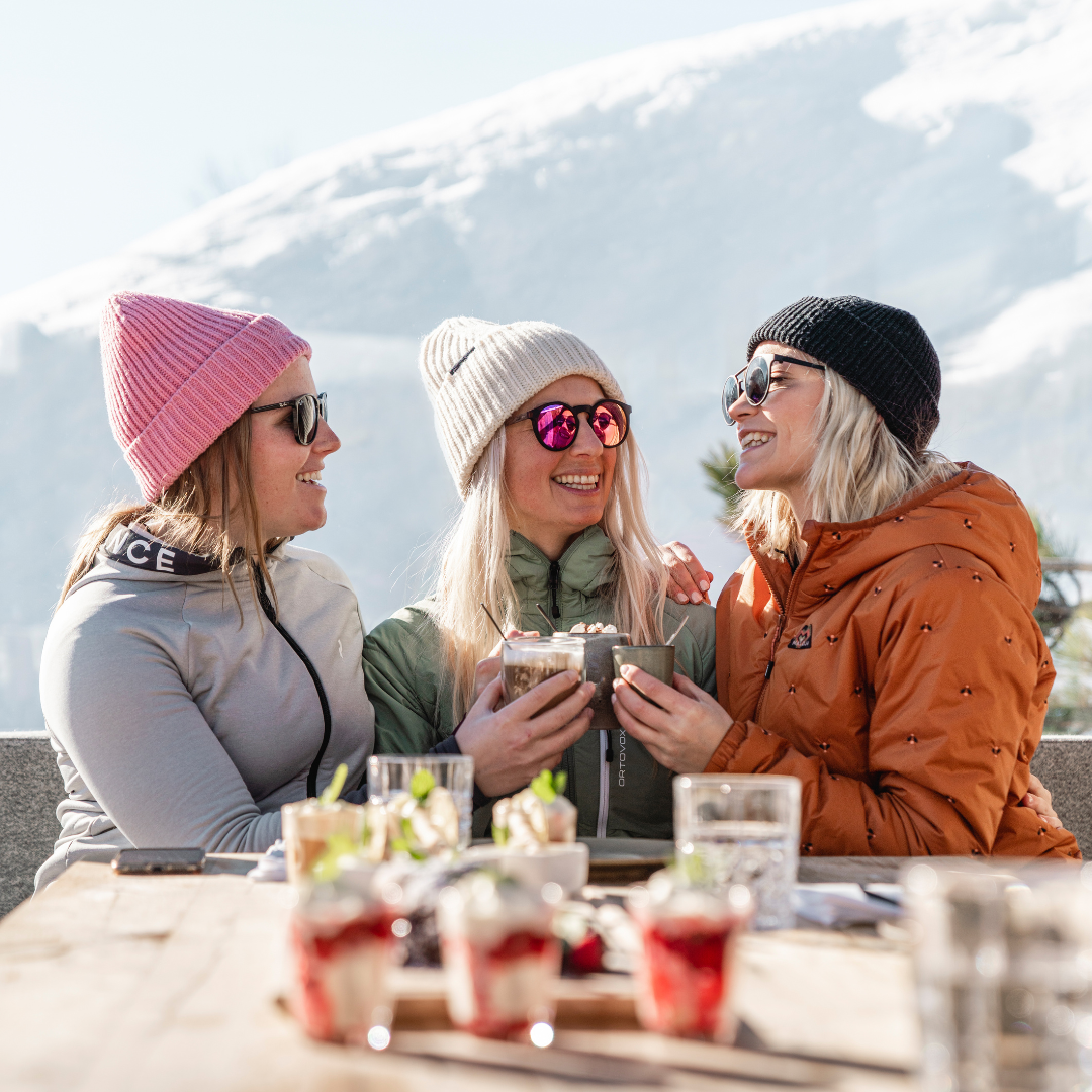 Tre personer i vintertøj nyder varme drikke og desserter ved et udendørs bord med sneklædte bjerge i baggrunden. De omfavner afterski-stemningen, smiler og bærer solbriller og strikhuer, hvilket skaber en munter og afslappet atmosfære.
