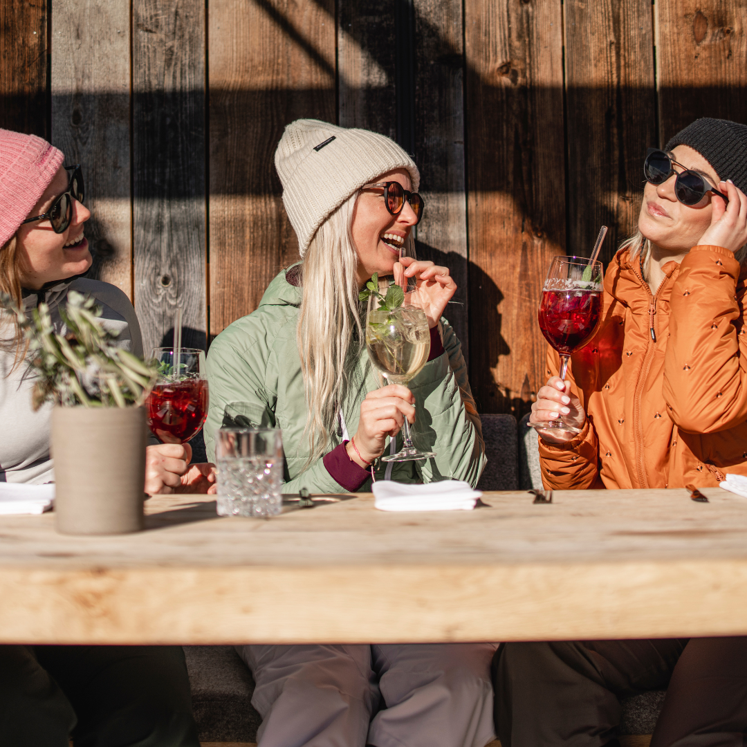 Tre kvinder i vintertøj sidder ved et træbord udendørs, nyder farverige drinks og griner efter en spændende dag med afterski. To bruger solbriller og huer, mens den tredje justerer sine solbriller. En potteplante og menuer står på bordet.