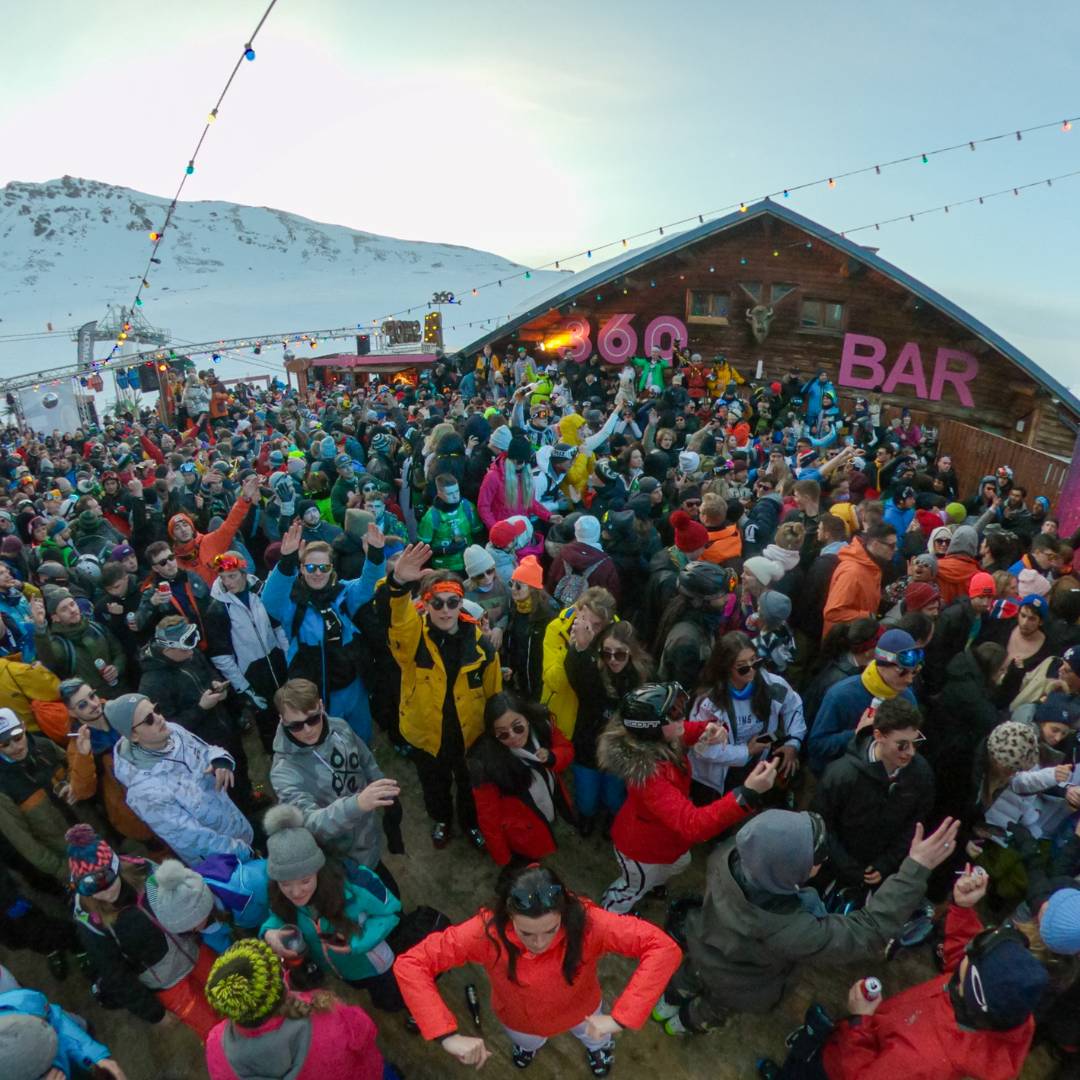 Et livligt publikum på en udendørs bar i sneklædte bjergomgivelser, der fejrer Nytår på ski. Folk er klædt i farverigt vintertøj og nyder en festlig atmosfære med lys spændt ovenover. En træbygning med BAR i lyserøde bogstaver står stolt i baggrunden.