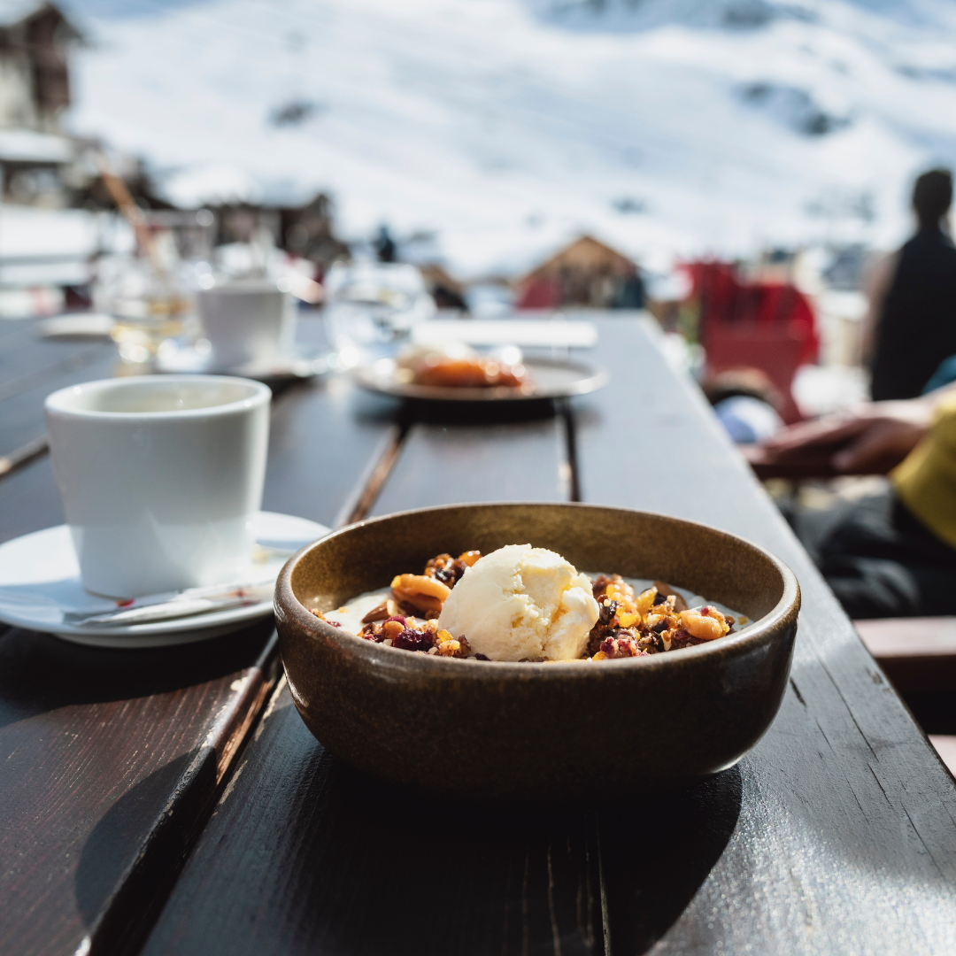 En skål dessert med is og nødder står på et træbord udendørs, omgivet af snedækkede bjerge, og danner den perfekte scene for Nytår på ski. En kop kaffe og en sløret skikkelse i vintertøj er synlige i baggrunden.