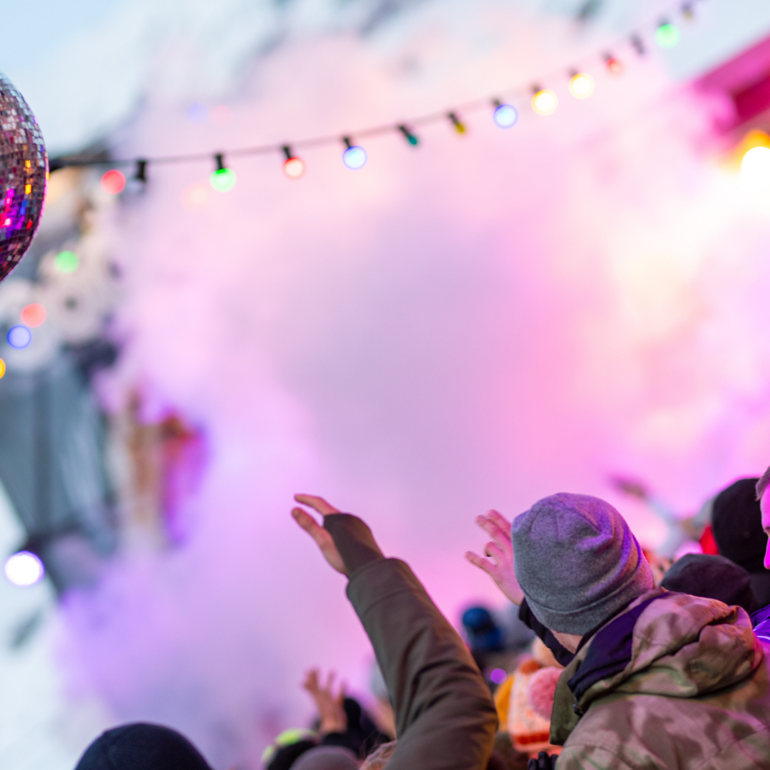 Et livligt publikum klædt i vintertøj nyder en udendørs fest, der minder om Nytår på ski. Klare, farverige lys glimter i den tågede atmosfære, mens en diskokugle afspejler den pulserende scene ovenfor.