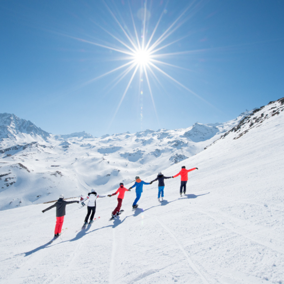 En gruppe på fem personer nyder Nytår på ski og står på ski ned ad en sneklædt bjergskråning under en skarp, skinnende sol. De holder hinanden i hånden, klædt i levende skiudstyr. Snedækkede tinder står majestætiske i baggrunden mod en klar blå himmel.