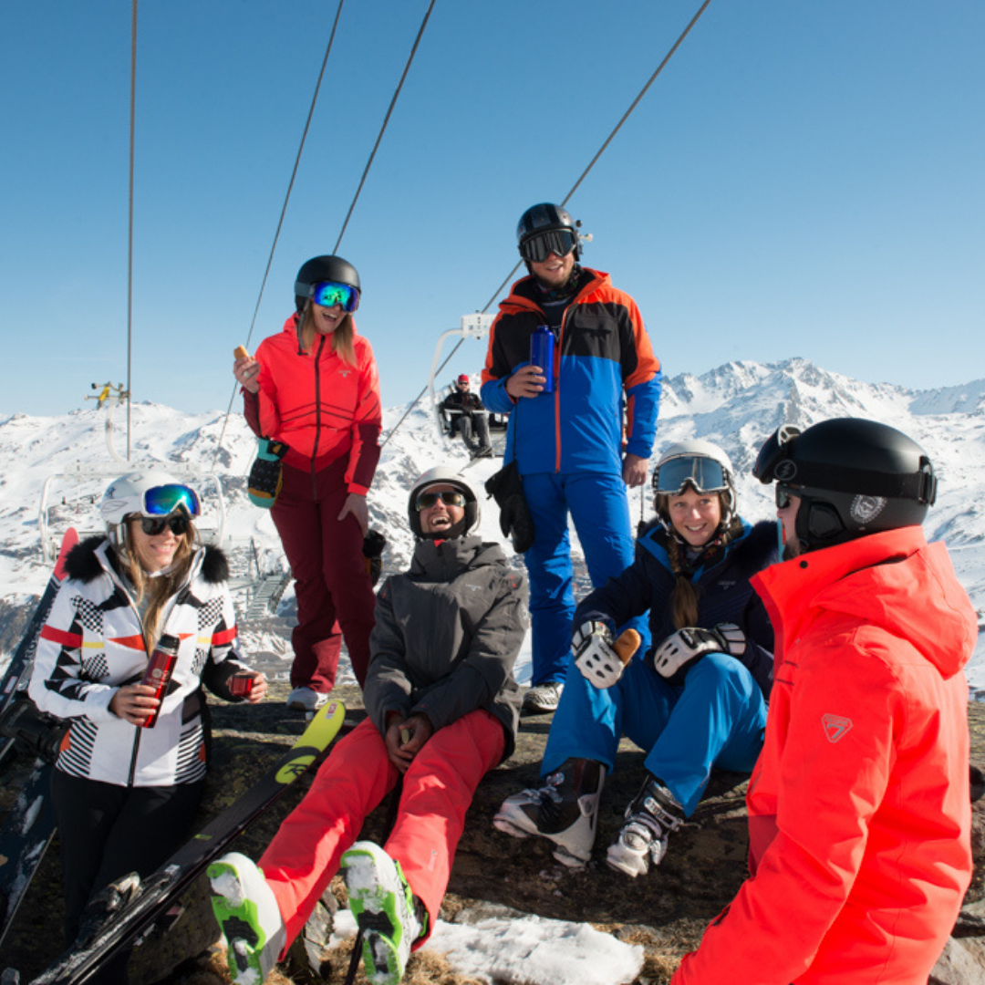 En gruppe på seks skiløbere i farverigt udstyr sidder og står på en sneklædt bjergskråning under en klar blå himmel. For at fejre Nytår på ski, er de i nærheden af en skilift, nyder en pause med drinks, omgivet af naturskønne snedækkede tinder.