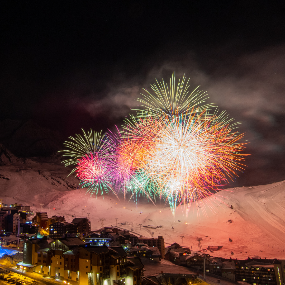 Farverigt fyrværkeri lyser nattehimlen op over et sneklædt bjergskisportssted og skaber den perfekte scene til en Nytår på ski-fest. Forskellige nuancer af rød, grøn og lilla eksploderer ovenover. Nedenfor er feriestedets bygninger oplyst i kontrast til det uberørte vinterlandskab.