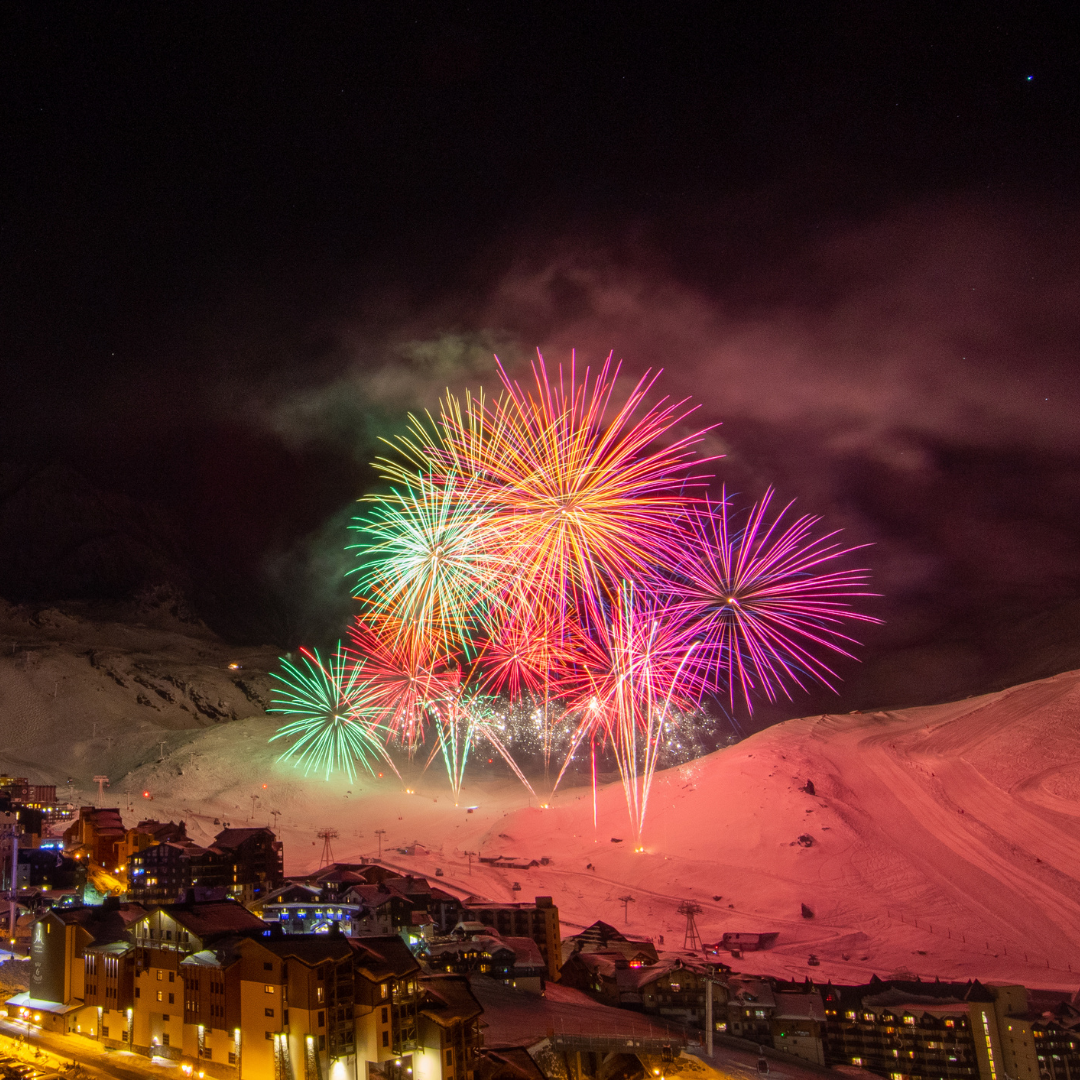 Et levende fyrværkeri lyser nattehimlen op over et sneklædt bjerglandskab under Nytår på ski. Lyse farver, inklusive pink, grøn og gul, oplyser området. Nedenfor ligger en by med oplyste bygninger i den hvide, vinterlige scene.
