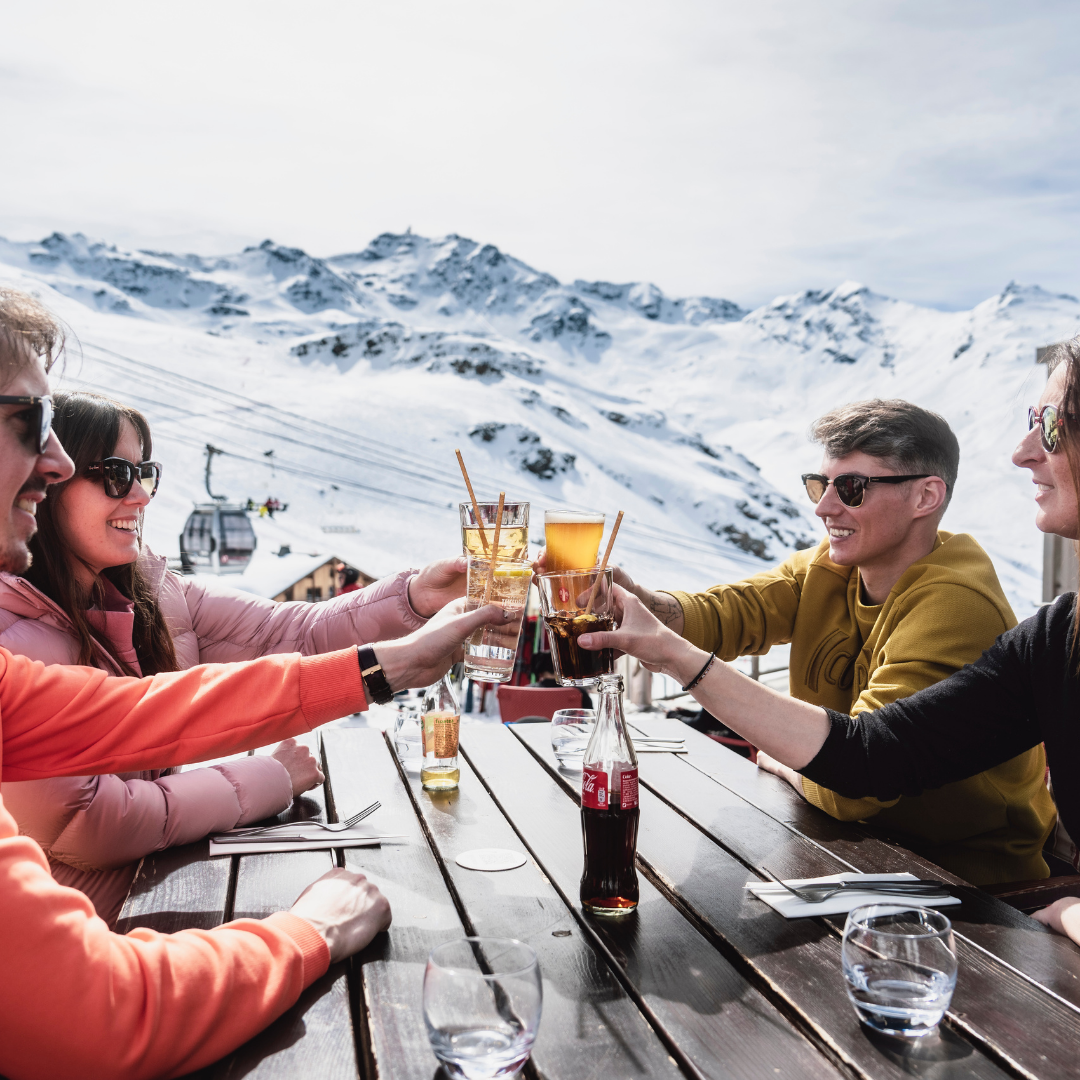 Fire personer skåler med drinks ved et udendørs træbord mod en sneklædt bjergkulisse, der fejrer nytår på ski. Klædt i vintertøj og solbriller nyder de den solrige dag, mens en svævebane glider forbi i det fjerne.