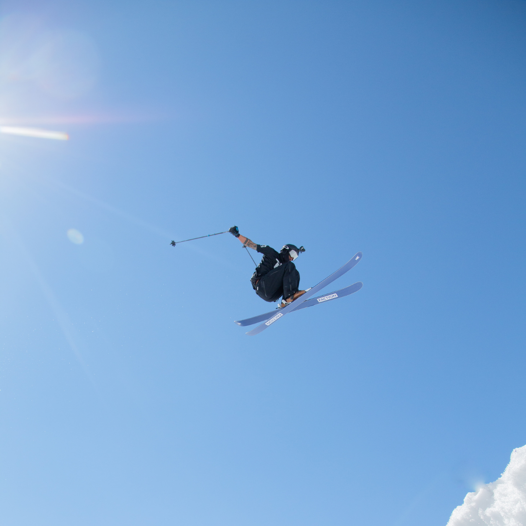 En skiløber fejrer Nytår på ski med et højtflyvende trick mod en klar blå himmel. Solen kaster et skarpt skær øverst til venstre, mens atleten svæver midt i springet, griber om skistave og iført sig beskyttelseshjelm og vinterudstyr.