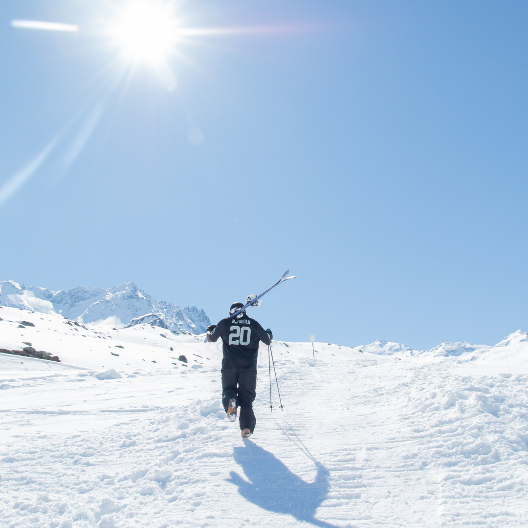 En person i en sportstrøje med tallet 20 bærer ski over skulderen og fejrer Nytår på ski, mens han går op ad bakke i et snelandskab under en strålende sol. Snedækkede bjerge rejser sig majestætisk i baggrunden mod en klar blå himmel.