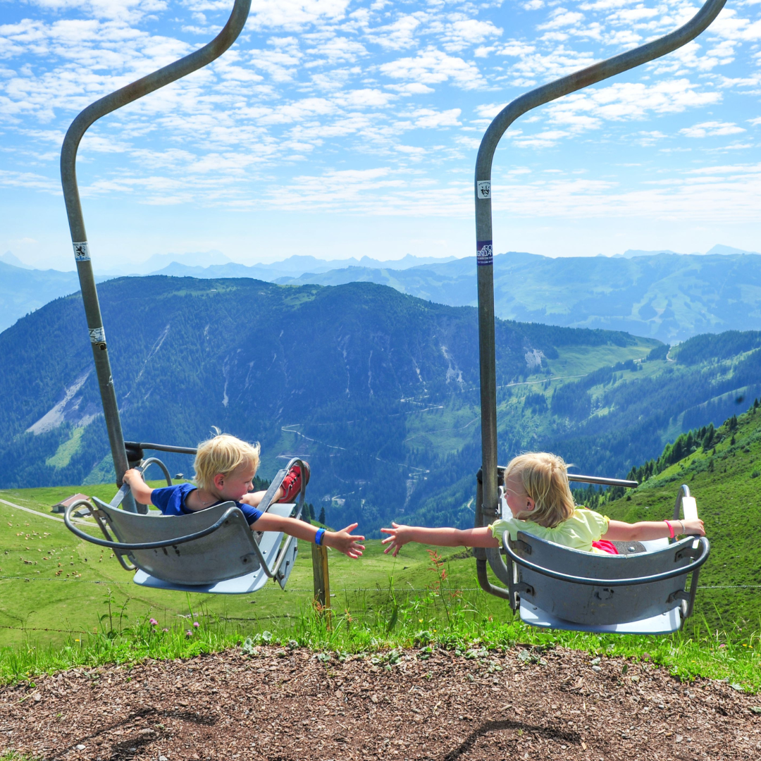 To børn i stoleliften rækker ud mod hinanden, mod det naturskønne bjerglandskab i Westendorf. Den delvist overskyede himmel og de pulserende grønne græsklædte bakker nedenfor fanger essensen af et perfekt sommerferieeventyr.