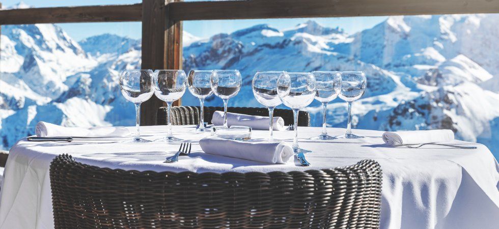 Ett runt bord dukat med vinglas, bestick och vita servetter på en vit duk, omgivet av korgstolar på en terrass med utsikt över snöiga berg - perfekt för ett skiferie i Frankrike.