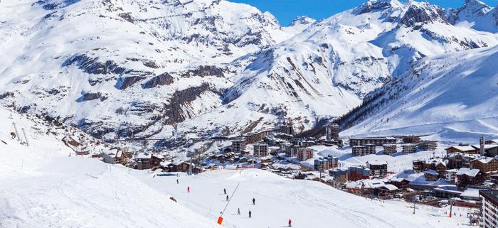 Ett snöigt bergslandskap med skidåkare i skidbackar, med utsikt över en stad som ligger inbäddad bland de snötäckta topparna under en klarblå himmel - perfekt för en skiferie i Frankrike.