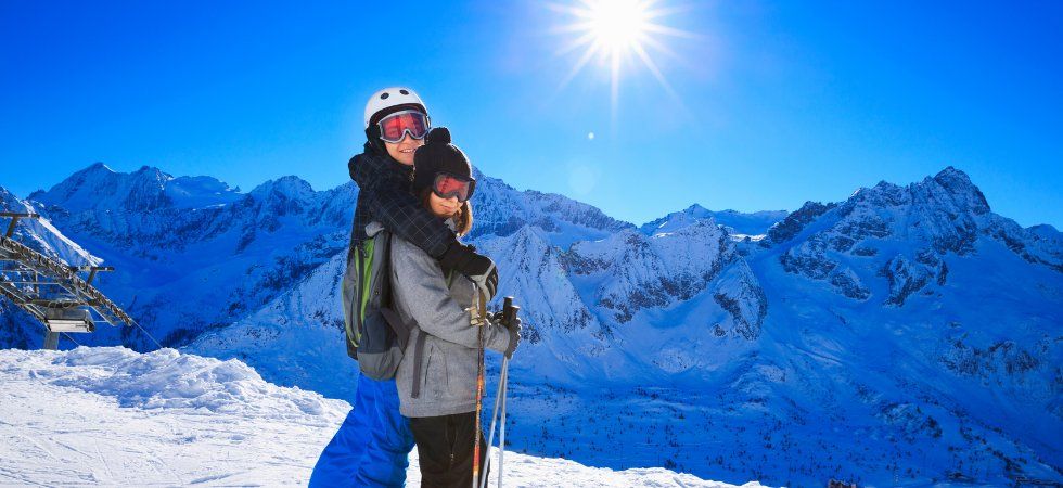 Två personer klädda i vinterkläder omfamnar varandra när de står på en snöig bergssluttning under sin skiferie i Frankrig, med skidstavar i handen. Snötäckta berg och en strålande sol mot en klarblå himmel kompletterar den fantastiska bakgrunden.