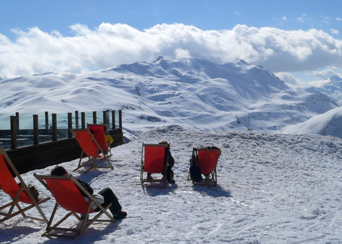 Folk kopplar av på röda solstolar på en snöig bergssluttning i Westendorf och njuter av den soliga dagen. Snötäckta toppar sträcker sig i bakgrunden under en blå himmel med spridda moln. Ett trästaket kantar den vänstra sidan.