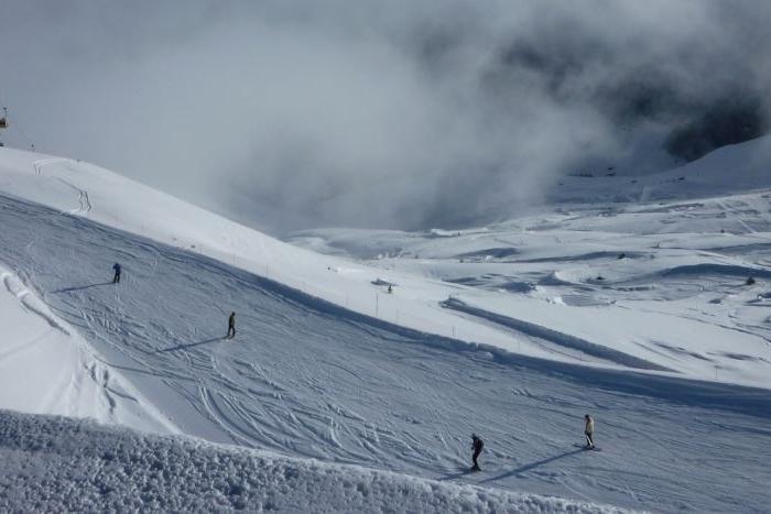Skidåkare glider nedför en snöig bergssluttning under molnig himmel, med liftstolar synliga till vänster och snötäckta kullar som sträcker sig i fjärran.