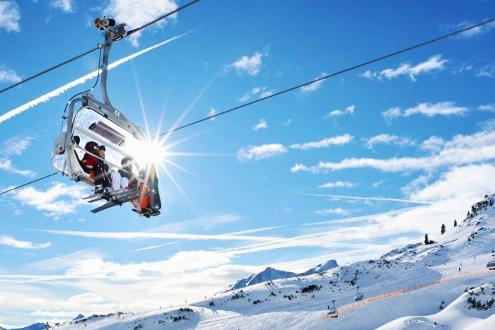 Skidåkare åker stollift uppför ett snöigt berg under en klarblå himmel med sol som skiner igenom, omgiven av snötäckta backar och avlägsna toppar.