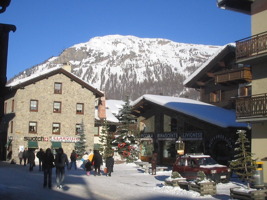 En snöig byscen i Saalbach fångar människor som går på en gata kantad av stenbyggnader och träbalkonger. Snötäckta berg, perfekta för en skidsemester, reser sig under en klarblå himmel. Ett rött fordon och små träd ger charm till denna idylliska skidresa.