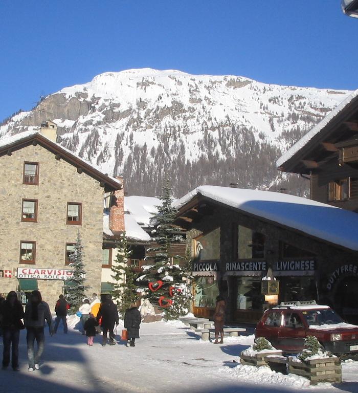 En snöig byscen i Saalbach fångar människor som går på en gata kantad av stenbyggnader och träbalkonger. Snötäckta berg, perfekta för en skidsemester, reser sig under en klarblå himmel. Ett rött fordon och små träd ger charm till denna idylliska skidresa.