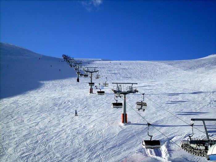 En snöig skidbacke i Saalbach med en skidlift uppför centrum. Några skidåkare navigerar nerför backen under en klarblå himmel. Liften har flera stolar, och spår från tidigare skidåkare kan ses i snön, vilket gör den till en perfekt plats för en minnesvärd skidsemester.