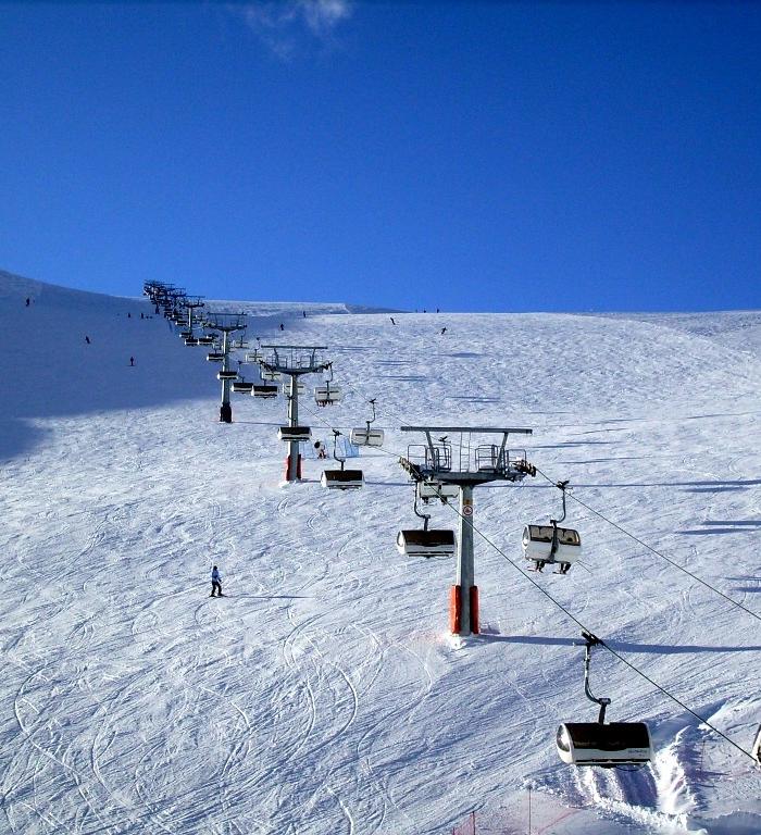 En snöig skidbacke i Saalbach med en skidlift uppför centrum. Några skidåkare navigerar nerför backen under en klarblå himmel. Liften har flera stolar, och spår från tidigare skidåkare kan ses i snön, vilket gör den till en perfekt plats för en minnesvärd skidsemester.