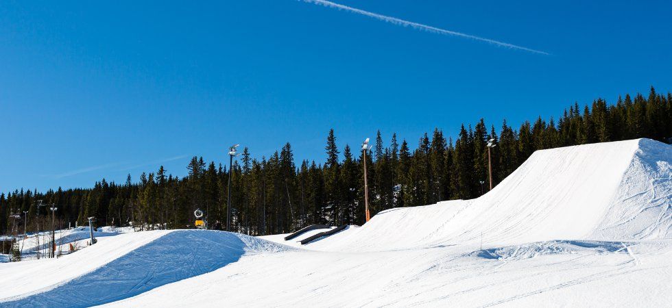 En snöig terrängpark med flera skid- och snowboardhopp under en klarblå himmel, omgiven av höga vintergröna träd - perfekt för en skiferie i Norge.