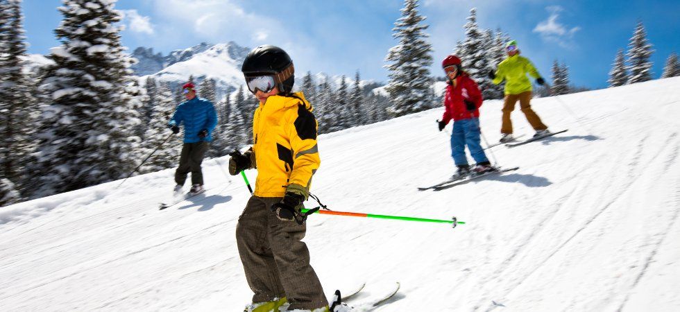 Fyra personer i färgglada vinterkläder åker skidor i Norge, utför en snöig bergssluttning, omgivna av snötäckta träd och berg under en ljusblå himmel.