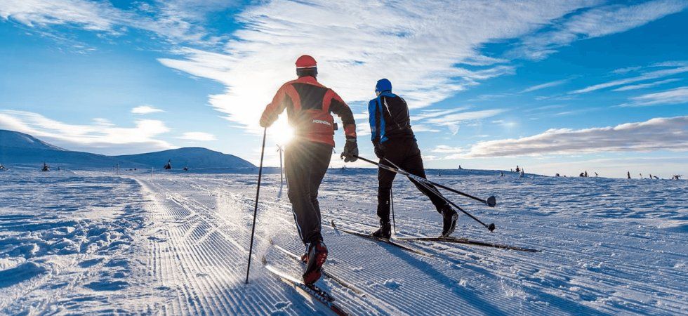 Två personer njuter av en skiferie i Norge, längdskidåkning i ett snöigt landskap under en klarblå himmel, med solen lågt i horisonten och berg som reser sig i bakgrunden.