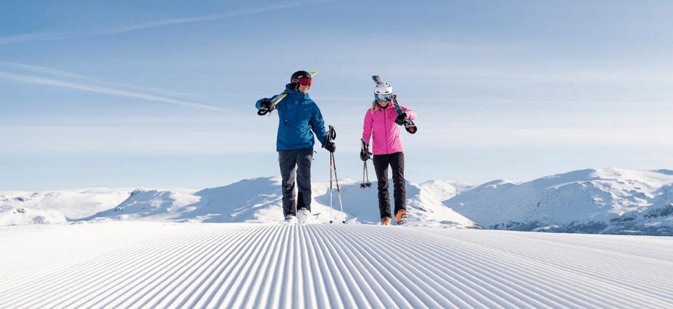 Två personer klädda i vinterkläder går på en nypreparerad snöig backe med skidor över axlarna - perfekt för en skiferie i Norge - med snötäckta berg och en klarblå himmel i bakgrunden.
