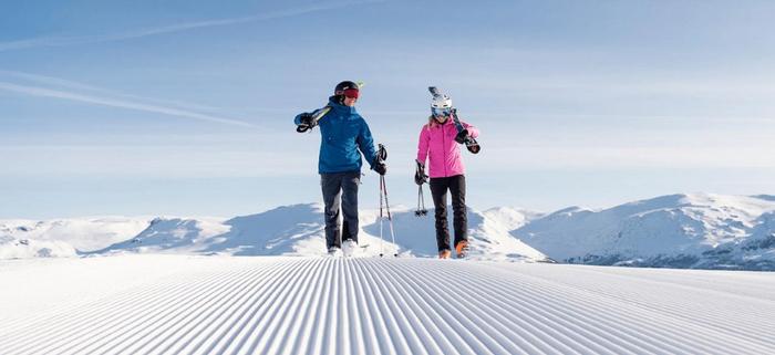 Två personer klädda i vinterkläder går på en nypreparerad snöig backe med skidor över axlarna - perfekt för en skiferie i Norge - med snötäckta berg och en klarblå himmel i bakgrunden.