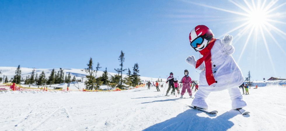 En person i snögubbedräkt med röd halsduk och hjälm åker snowboard nedför en solig sluttning under en skiferie i Norge, medan skidåkare och barn i vinterkläder står i bakgrunden under en klarblå himmel.