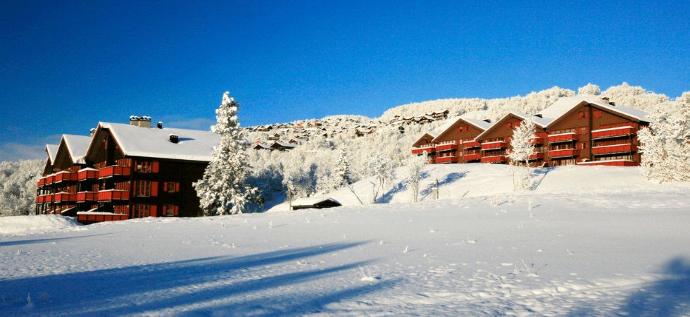 Röda trästugor med snötäckta tak ligger på en snöig sluttning, omgivna av snötyngda träd under en klarblå himmel - en idyllisk skiferie i Norge, där charmiga hus pryder det frostiga vinterlandskapet.