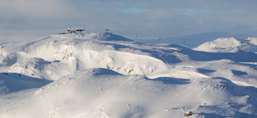 Snötäckt bergslandskap med mjuka sluttningar, en skidlift som går över kullarna och en liten byggnad nära toppen under en delvis molnig himmel - perfekt för en skiferie i Norge.
