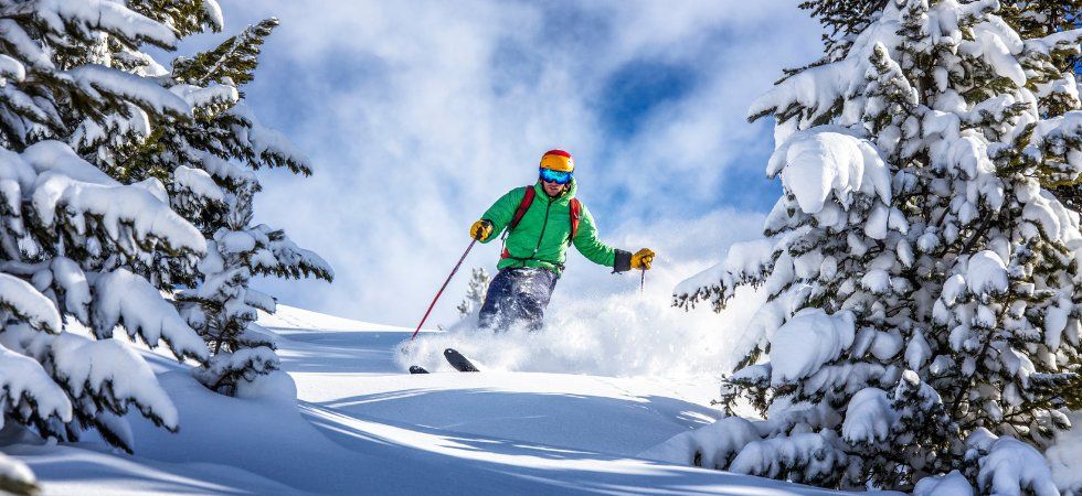 En skidåkare i grön jacka, blå byxor och färgglad mössa njuter av en skiferie i Østrig och glider nerför backen genom nysnö mellan tallar en solig vinterdag.