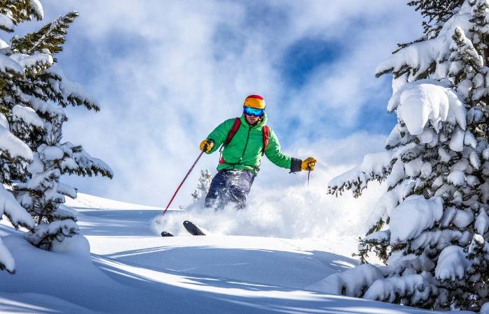 En skidåkare i grön jacka, blå byxor och färgglad mössa njuter av en skiferie i Østrig och glider nerför backen genom nysnö mellan tallar en solig vinterdag.