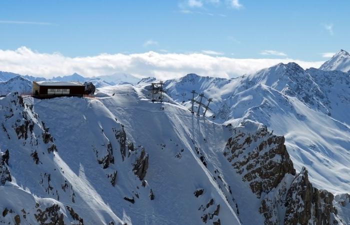 En snöklädd bergstopp med en byggnad och skidlift på toppen, perfekt för skiferie i Østrig, omgiven av karga, snötäckta berg under en ljusblå himmel med spridda moln.