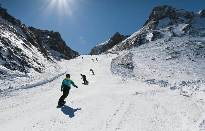 Människor åker snowboard och skidor nedför en bred, snöig bergssluttning på en solig skiferie i Österrike, omgiven av klippiga toppar under en klarblå himmel.