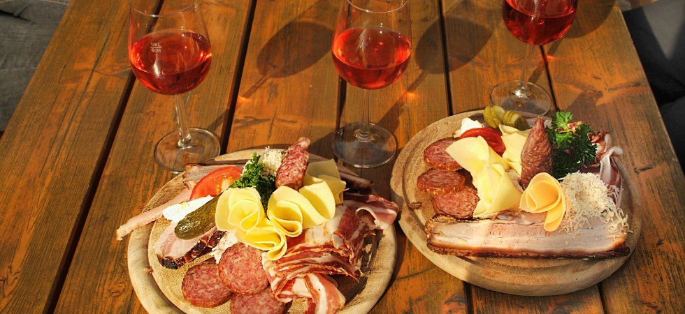 Två trätallrikar med olika sorters kallskuret, ostar, pickles och bröd står uppdukade på ett träbord tillsammans med tre glas rosévin. Solljuset framhäver den livfulla uppläggningen - perfekt efter en dag på skiferie i Østrig.